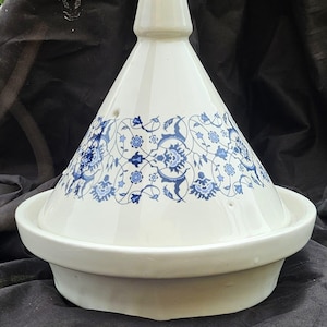 Puede incluir: Tajine de cerámica blanca con tapa cónica y base ancha. La tapa está decorada con un estampado floral azul. El tajine está diseñado para cocinar a fuego lento, un recipiente de cocina marroquí tradicional.