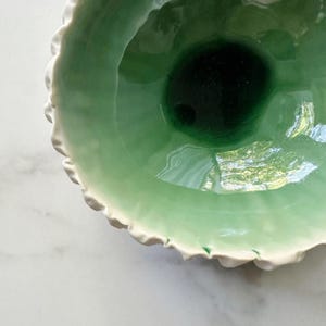 Könnte beinhalten: Eine Keramikschale mit glänzender Oberfläche, die einen Farbverlauf von hellem Minzgrün zu einem tiefen Waldgrün in der Mitte aufweist. Der Rand ist mit unregelmäßigen, gewellten weißen Kanten strukturiert, die einen Kontrast zum glatten Inneren bilden.
