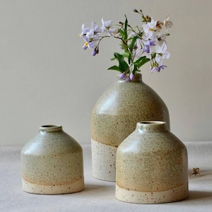 Könnte beinhalten: Drei Keramikvasen in verschiedenen Größen mit einer gesprenkelten, erdig-grünen Glasur und einem cremefarbenen Sockel. Die größte Vase enthält zarte lila und weiße Blumen, die der Anordnung Farbe verleihen.