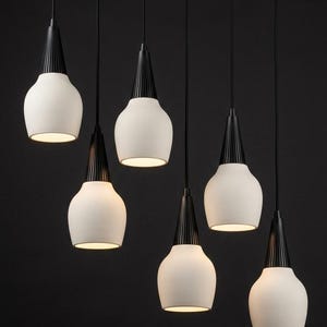 May include: Six pendant lights with white, rounded shades and black, cone-shaped tops hang against a dark background. The lights are evenly spaced and illuminated, creating a modern and minimalist aesthetic.