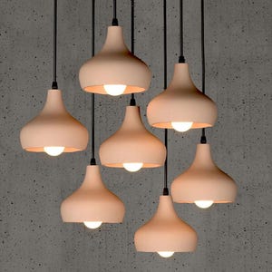 May include: Seven peach-coloured pendant lights hang from black cords against a grey textured wall. Each light has a rounded, bell-shaped shade with a bright white bulb, creating a warm glow. The lights are a modern home decor item.