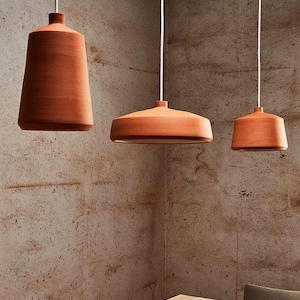 Op de afbeelding: Drie terracotta hanglampen van verschillende vormen hangen aan witte snoeren. De lampen hangen boven een houten tafel. De grootste lamp heeft een conische vorm, terwijl de andere ronder zijn.