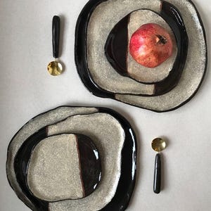 Pode incluir: Dois conjuntos de pratos decorativos com um design preto e prateado, cada um com uma pequena colher dourada e um cabo preto. Um conjunto tem uma romã vermelha. Os pratos têm uma forma orgânica e uma estética moderna.