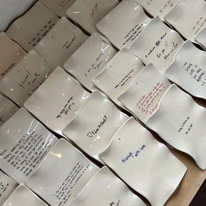 Pode incluir: Uma coleção de pratos de cerâmica branco-sujo com bordas onduladas, cada um com mensagens manuscritas em várias cores. Frases como "Eu te amo" e outros sentimentos são visíveis. Os pratos estão dispostos em um padrão de grade.