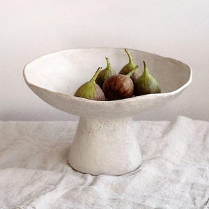 May include: A white ceramic pedestal bowl holding fresh figs. The bowl features a wide, shallow dish and a cylindrical base. The figs are green and brown. The bowl is placed on a textured, off-white linen cloth.