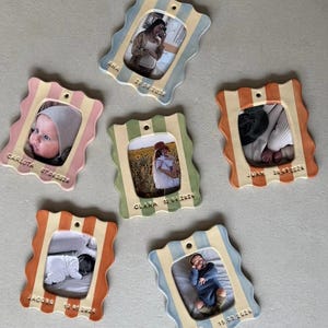Op de afbeelding: Zes keramische fotolijsten met golvende randen, elk met een andere foto. De lijsten zijn in verschillende kleuren, waaronder roze, blauw, groen en oranje, met strepenpatronen. Elke lijst heeft een naam en een datum.