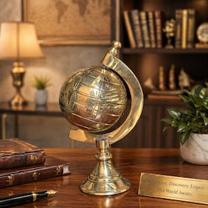 Könnte beinhalten: Ein Globus aus Messing mit gravierten Kontinenten und Ozeanen. Der Globus steht auf einem Holzschreibtisch mit Stift, Büchern und einem Schild mit der Aufschrift "Adventure. Discovery. Legacy. The World Awaits."