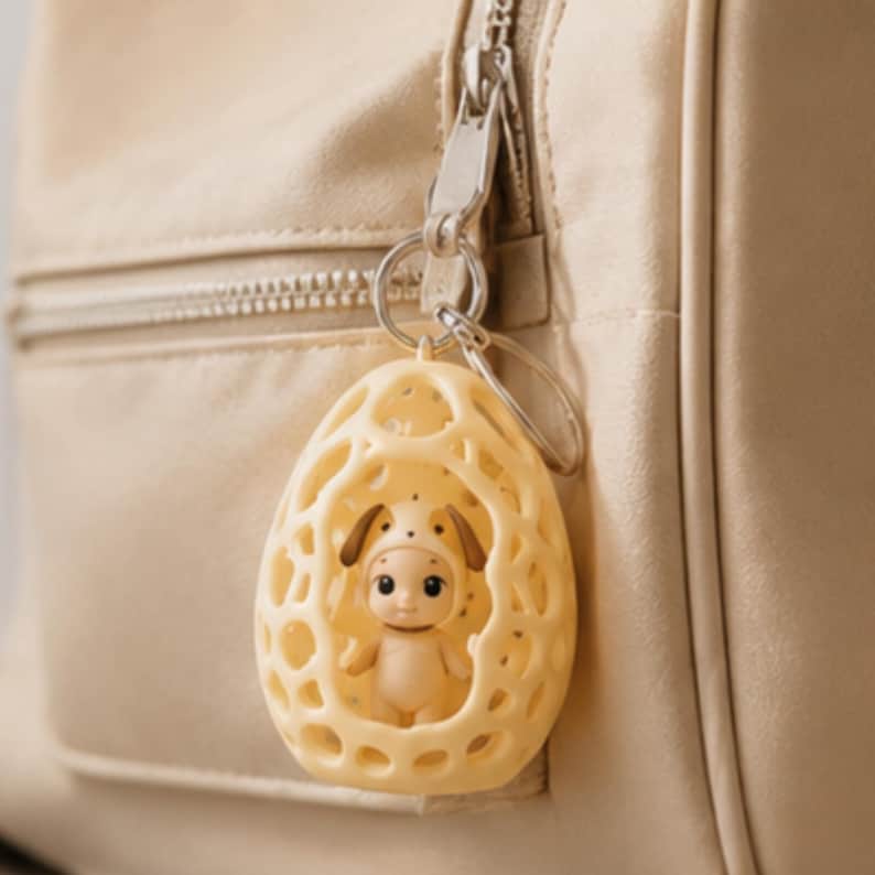 K&ouml;nnte beinhalten: Ein gelber Schl&uuml;sselanh&auml;nger mit einer kleinen Figur in einem dekorativen, eif&ouml;rmigen K&auml;fig. Die Figur tr&auml;gt ein Hundekost&uuml;m. Der Schl&uuml;sselanh&auml;nger ist an einem silbernen Ring befestigt und h&auml;ngt vor einer beigen Tasche.