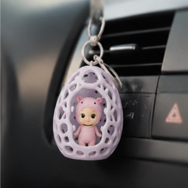 K&ouml;nnte beinhalten: Ein helllila, eif&ouml;rmiger Schl&uuml;sselanh&auml;nger mit Gittermuster, der eine kleine rosa Figur in einem Schweinekost&uuml;m h&auml;lt. Der Schl&uuml;sselanh&auml;nger h&auml;ngt vor dem Armaturenbrett eines Autos.