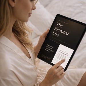 May include: A person holding a black tablet displaying the text "The Elevated Life" in white. The tablet screen also shows additional text and a white background. The person is wearing a cream-colored robe.