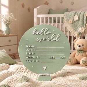 以下が含まれることがあります： 「hello world」の文字が白で書かれた、丸いセージグリーンのベビーアナウンスメントサイン。赤ちゃんの名前、日付、時間、体重（ポンド、オンス）、身長を書き込むスペースがあります。下部に小さなハートがあります。