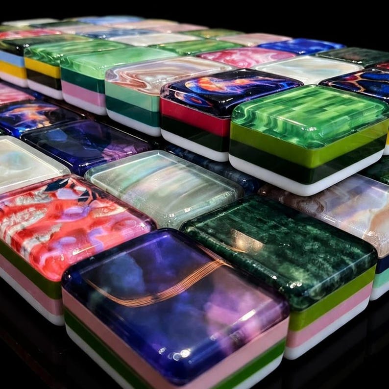 May include: A collection of rectangular tiles featuring diverse marbled and solid colour patterns. Each tile has a glossy, smooth surface with layered edges in various colours including pink, green, white, and black, arranged in a grid-like formation.