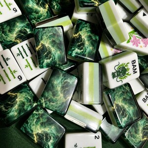 Peut inclure: Une collection de tuiles de mah-jong comprenant des tuiles blanches avec des motifs de bambou et de dragon verts, ainsi que des tuiles au motif de n&eacute;buleuse vert fonc&eacute; et dor&eacute;. Certaines tuiles portent le texte BAM et des chiffres en vert.