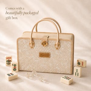 May include: A rectangular beige carrying case with two top handles and a central gold-toned clover-shaped clasp. The front features a textured pattern and a rectangular patch with the text Thank you. Several mahjong tiles and dice are nearby.