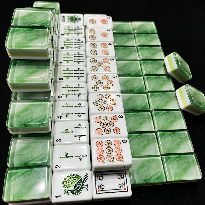 May include: A set of rectangular mahjong tiles featuring a white base with green marbled backs. The tiles display various traditional symbols, including bamboo stalks, circular patterns, a peacock illustration, and floral motifs in green and orange.