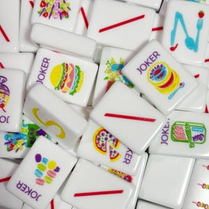May include: A collection of white rectangular mahjong tiles featuring colorful printed graphics of food items like burgers, pizza, and macarons, alongside tiles marked with the word JOKER in purple text and others displaying simple red diagonal lines.