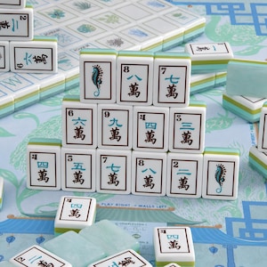 May include: A set of white mahjong tiles with light blue and green accents, featuring black Chinese characters and seahorse illustrations. The tiles are arranged in a pyramid formation on a light blue game board with printed text and patterns.