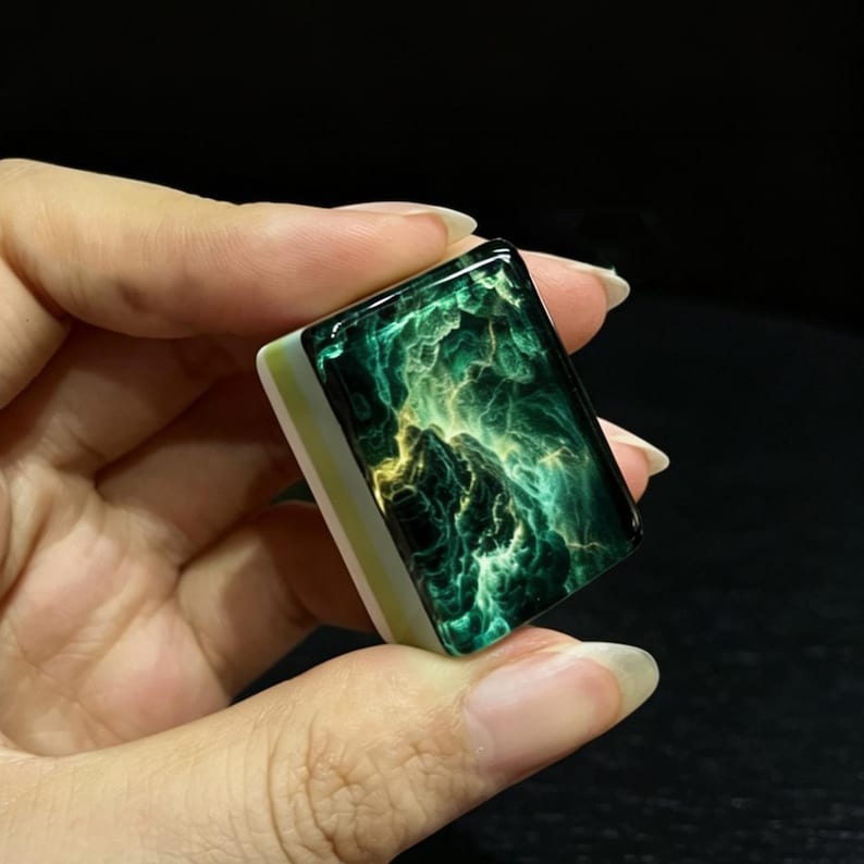 Peut inclure: Une tuile de mah-jong rectangulaire pr&eacute;sentant une surface brillante vert fonc&eacute; et noire avec des motifs tourbillonnants semblables &agrave; une n&eacute;buleuse dans des tons jaune vif et vert citron. La base est en mat&eacute;riau blanc opaque avec une fine rayure vert clair.