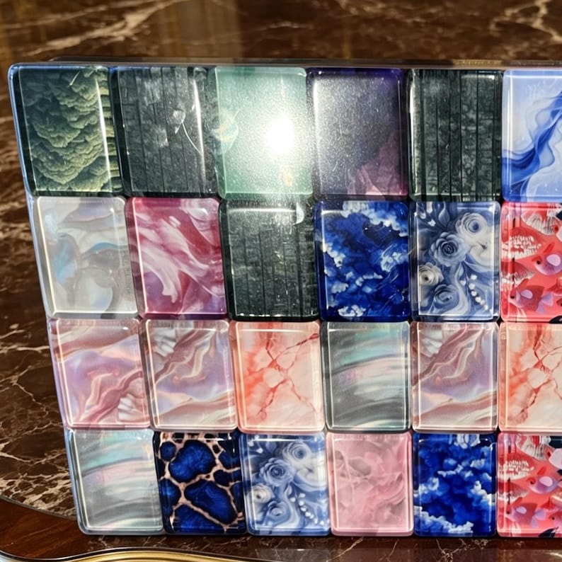 May include: A rectangular set of mahjong tiles arranged in a grid, featuring a variety of colourful designs including floral patterns, marbled textures, blue and pink swirls, dark green foliage, and animal print motifs on a glossy, smooth surface.