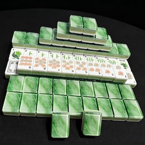 May include: A set of rectangular mahjong tiles arranged in rows and a pyramid stack. The tiles feature a white base with green marbled backs. Some tiles display orange circular patterns, green bamboo symbols, a peacock illustration, and the text BAM.