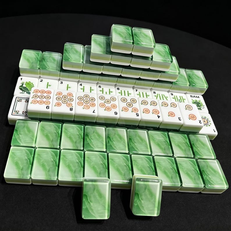 May include: A set of rectangular mahjong tiles arranged in rows and a pyramid stack. The tiles feature a white base with green marbled backs. Some tiles display orange circular patterns, green bamboo symbols, a peacock illustration, and the text BAM.