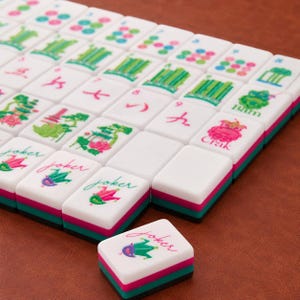 May include: A set of white Mahjong tiles with colorful illustrations including green bamboo, pink flowers, and joker icons. Each tile features a layered base with a black, pink, and teal stripe design. One tile is placed separately in the foreground.