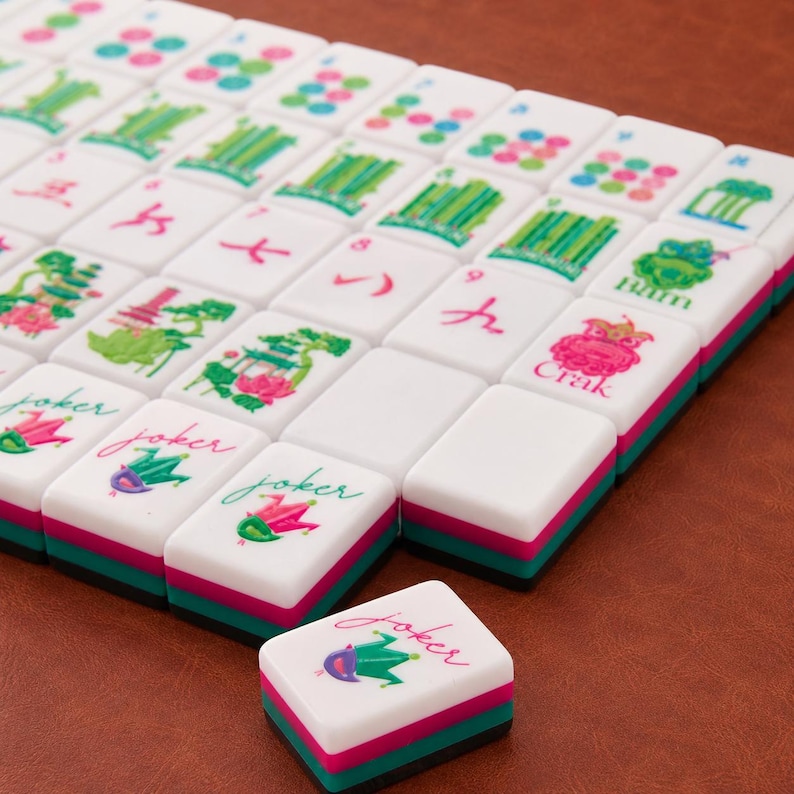 May include: A set of white Mahjong tiles with colorful illustrations including green bamboo, pink flowers, and joker icons. Each tile features a layered base with a black, pink, and teal stripe design. One tile is placed separately in the foreground.