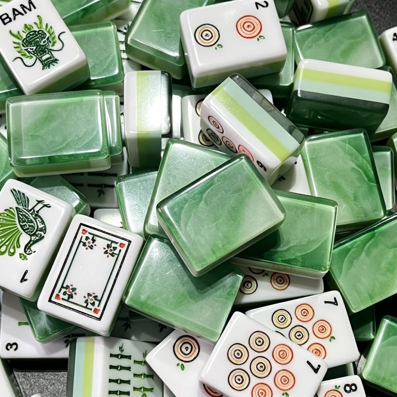 May include: A collection of mahjong tiles featuring a mix of white and marbled green surfaces. Some tiles display black ink illustrations of dragons, birds, and bamboo, while others show red and orange circular patterns and numerical markings.