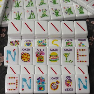 May include: A collection of white rectangular mahjong tiles featuring colorful illustrations of food, flowers, and utensils. Some tiles display the word JOKER in purple text, while others show red diagonal lines or various graphic designs like a burger, pizza, and cocktails.