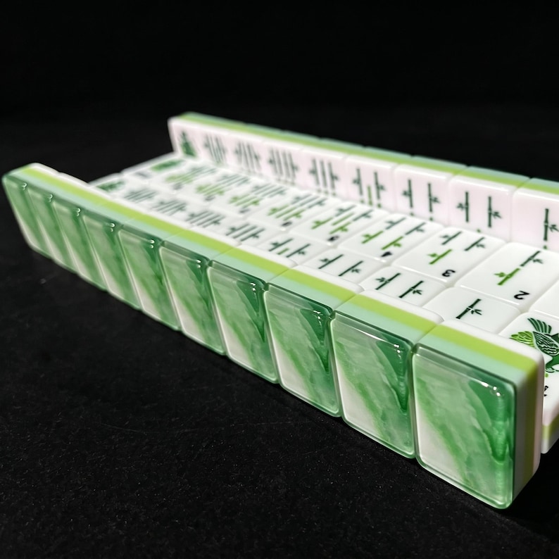 May include: A set of rectangular mahjong tiles arranged in a row on a green and white marbled rack. The tiles are white with green and black engraved symbols, including bamboo patterns and numbers, featuring a smooth, glossy finish on the front.