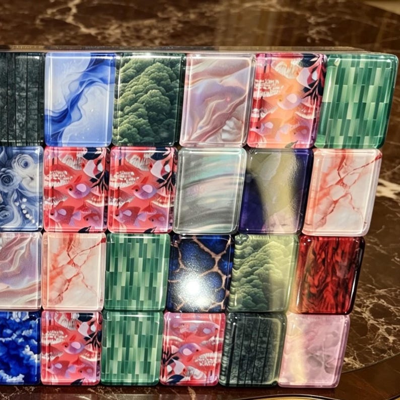May include: A rectangular grid of twenty-four small, glossy tiles arranged in four rows and six columns. Each tile features a distinct pattern, including swirling blue and white, green foliage, pink marble, orange floral, and red fish motifs.