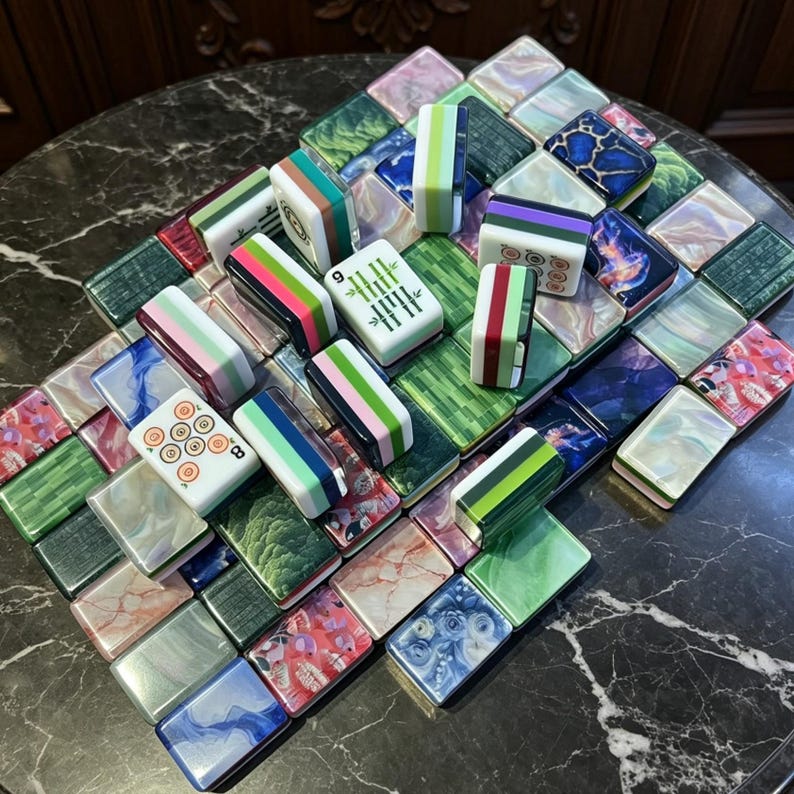 May include: A collection of rectangular mahjong tiles featuring a variety of patterns, including solid colours, marbled textures, floral prints, and traditional bamboo and circle markings, arranged in a scattered, overlapping formation.