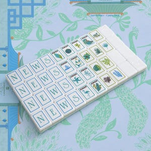 May include: A set of rectangular white mahjong tiles arranged in a grid. The tiles feature blue lettered tiles spelling NEWS, tiles labeled JOKER with various tropical illustrations like flamingos, starfish, and palm trees, and blank white tiles.
