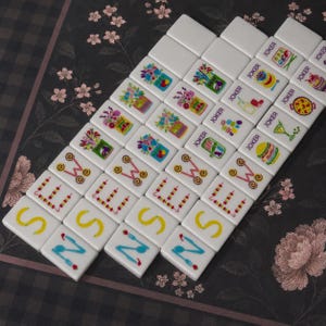May include: A set of white rectangular mahjong tiles arranged in a grid. The tiles feature colorful printed graphics including floral arrangements, food items, drinks, and the word JOKER, alongside letters S, E, W, and N with decorative accents.