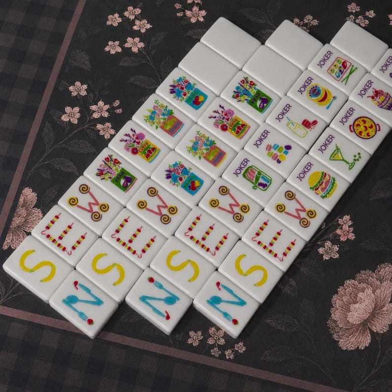 May include: A set of white rectangular mahjong tiles arranged in a grid. The tiles feature colorful printed graphics including floral arrangements, food items, drinks, and the word JOKER, alongside letters S, E, W, and N with decorative accents.