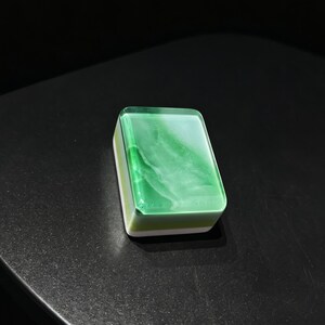 May include: A rectangular mahjong tile featuring a translucent green top layer with white marbled swirls. The base of the tile is constructed from a solid white material, creating a distinct layered appearance with smooth, rounded edges and corners.