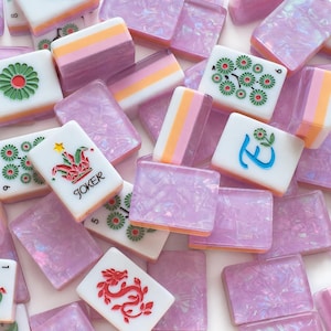 Peut inclure: Une collection de tuiles de mah-jong rectangulaires présentant un mélange de faces blanches avec des motifs imprimés colorés, incluant un joker, un dragon rouge et des motifs floraux, associés à des dos translucides, lavande et rose pailletés.