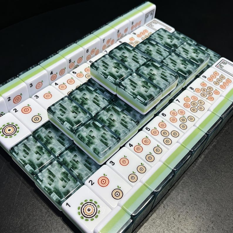 May include: A set of rectangular mahjong tiles arranged in a grid. Some tiles feature white surfaces with orange and green circular target-like symbols and black numbers, while others have a translucent, marbled dark green and white pattern.
