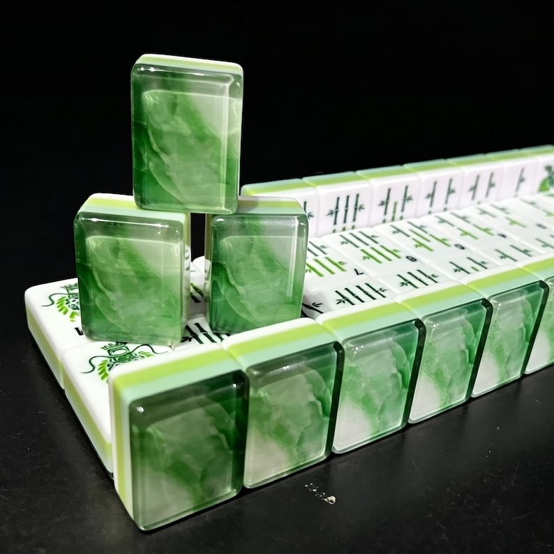 May include: A set of rectangular mahjong tiles featuring a marbled green and white pattern on the back and white faces with green engraved symbols, including bamboo stalks and dragon motifs, arranged in a stacked formation on a dark surface.