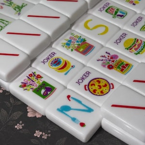 May include: A collection of white rectangular Mahjong tiles featuring colorful illustrations including a yellow letter S, a burger, a pizza, a spoon and fork, and various floral arrangements. Several tiles are marked with the word JOKER.