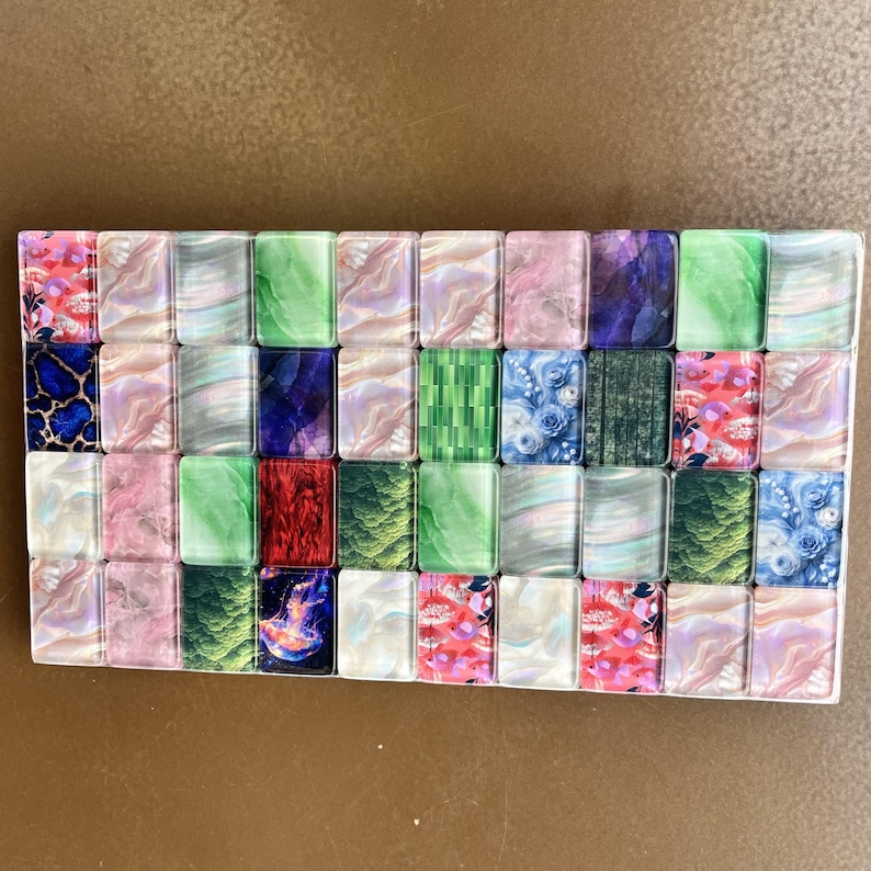 May include: A rectangular set of forty small, smooth, glass-like tiles arranged in a grid. Each tile features a unique design, including marbled patterns in pink, green, and white, floral prints, deep blue textures, and a vibrant jellyfish graphic.