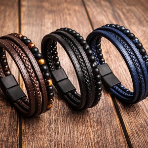 Peut inclure: Trois bracelets multicouches en marron, noir et bleu. Chaque bracelet présente une combinaison de cuir tressé, de cuir lisse et d'accents perlés. Les bracelets ont des fermoirs magnétiques et sont présentés sur une surface en bois.