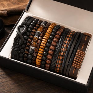 Op de afbeelding: Een set van meerdere armbanden in een zwarte doos, inclusief zwarte leren banden met een oneindigheidssymbool, diverse kralenstrengen in zwart, bruin en tijgeroog, en gevlochten leren banden in zwart en bruin met stikseldetails.