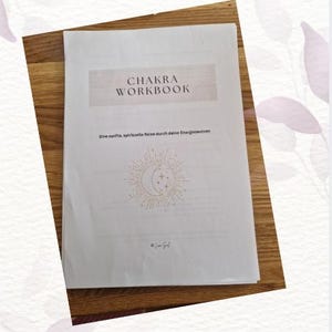 Könnte beinhalten: Ein weißes Arbeitsbuch mit dem Titel "CHAKRA WORKBOOK" mit goldenem Text und einer Sonne- und Mond-Illustration. Das Arbeitsbuch liegt auf einer Holzoberfläche. Der Text "Eine sanfte, spirituelle Reise durch deine Energiezentren" steht unter dem Titel.