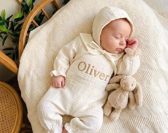 May include: A cream-colored long-sleeve romper with a matching hood, featuring the name Oliver embroidered in brown thread on the chest. The garment has ruffled cuffs and a tie closure at the neck, accompanied by a small beige plush rabbit toy.