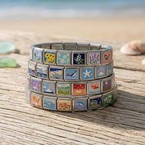 May include: Three stainless steel charm bracelets stacked on a wooden surface. Each bracelet features colorful square charms with various designs, including flowers, a whale, and a seashell. The background shows a beach scene with the ocean and sand.
