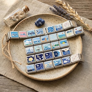 Peut inclure: Une collection de breloques carrées argentées avec des motifs émaillés colorés représentant des scènes marines, notamment des baleines, des voiliers et des couchers de soleil. Les breloques sont disposées sur une assiette en céramique tachetée.