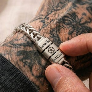 Pode incluir: Uma pulseira de corrente de metal prateado com três contas retangulares. As contas estão gravadas com o nome Ronaldo, um gráfico de uma bola de futebol e a data 06/11/2026. A pulseira tem um design de elos trançados e um acabamento metálico polido.