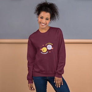 Puede incluir: Sudadera granate con el texto "Let's Spread Our Kindness" y un gráfico de dos planetas de dibujos animados. Un planeta es amarillo y el otro morado. La sudadera está hecha de un material suave.