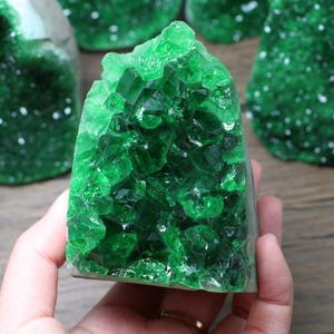 May include: A vibrant green crystal cluster with a sparkling, faceted surface. The crystal is a deep emerald color, with a rough, natural texture. Several similar crystals are visible in the background, creating a collection of natural gemstones.